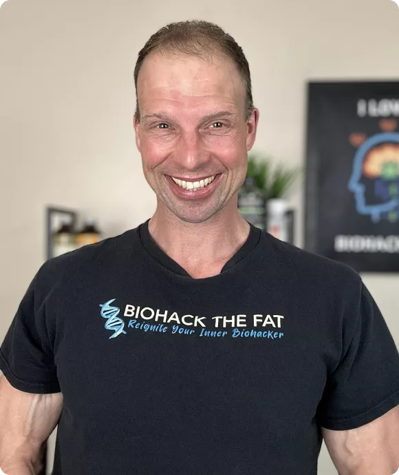 Trevor Folgering, founder of Biohack The Fat, smiling and wearing a branded shirt in his wellness and fitness workspace.